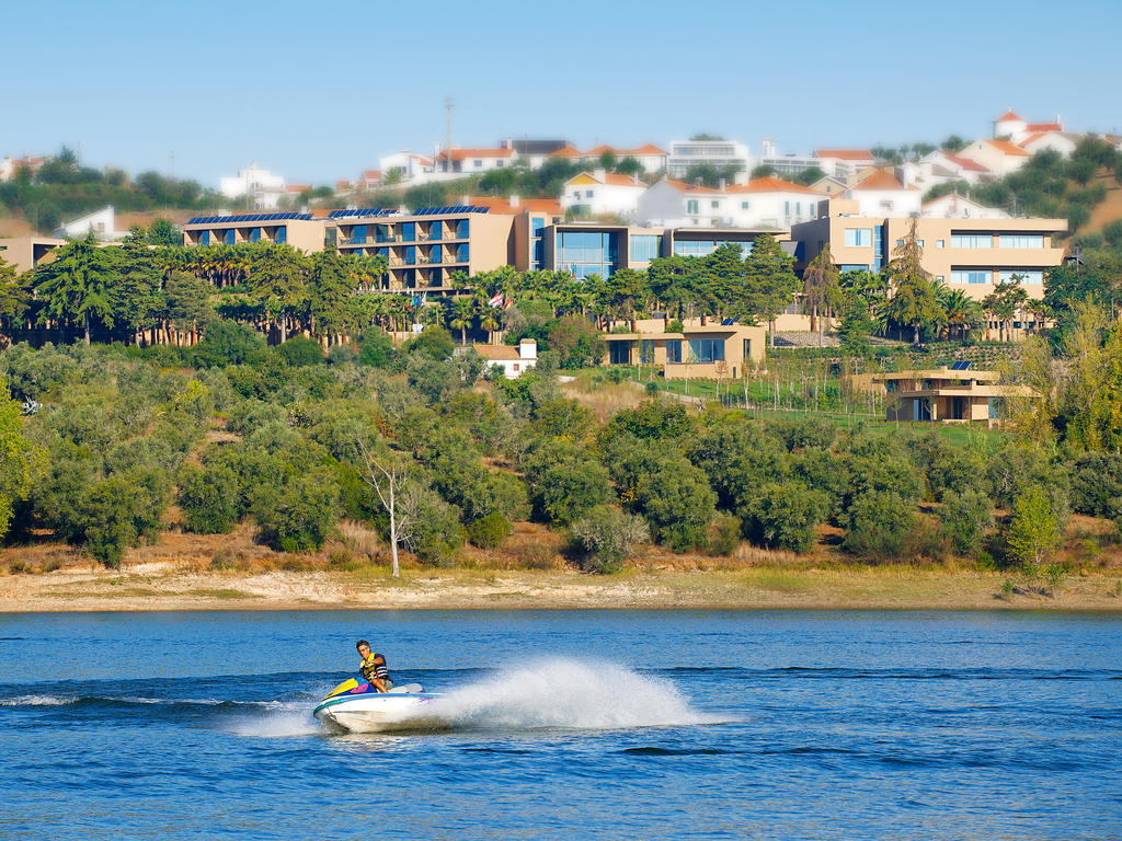 Ap Lago Montargil - Conference & Spa Extérieur photo The photo depicts a person riding a jet ski on a body of water. In the background, there are modern buildings and lush greenery, suggesting a scenic landscape, likely near a resort or upscale residential area. The clear blue sky and calm water enhanc