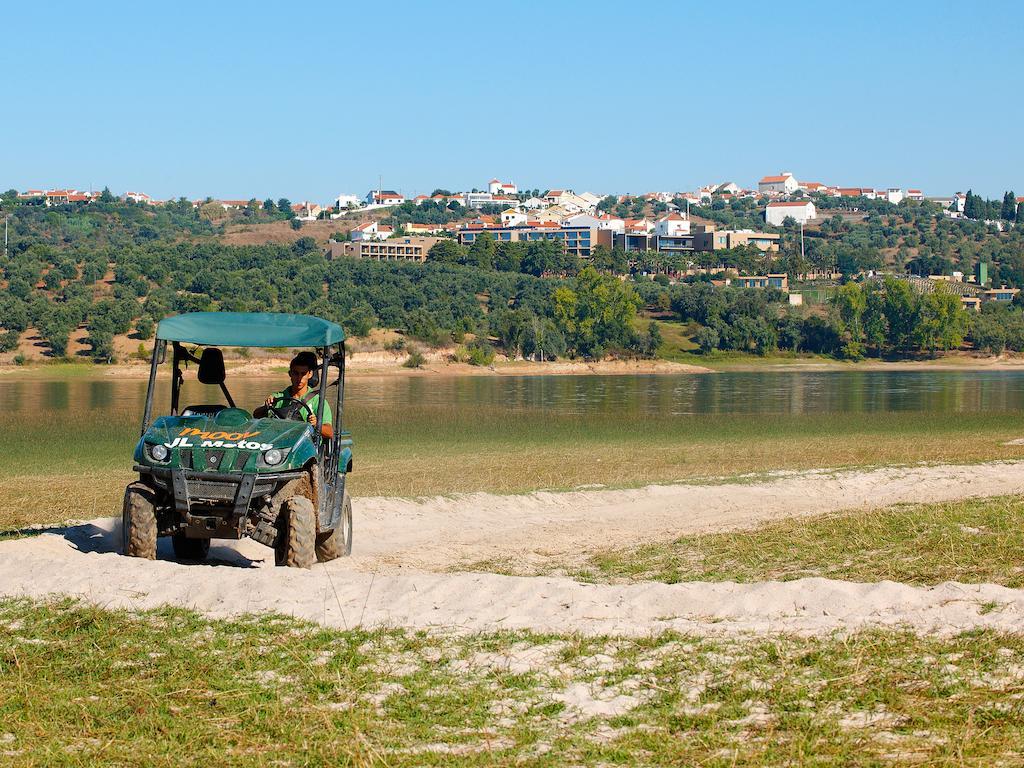 Ap Lago Montargil - Conference & Spa Extérieur photo The photo shows a small green all-terrain vehicle driving along a sandy path beside a body of water. In the background, there's a landscape with trees and rolling hills, along with a town or village visible on the hillside. The sky is clear, indicati