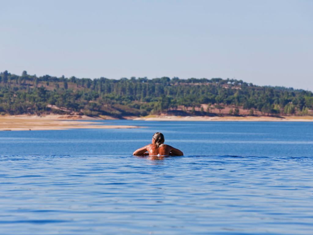 Ap Lago Montargil - Conference & Spa Extérieur photo The photo shows a person swimming in a calm body of water, likely a lake or a river. The swimmer is seen from behind and appears to be enjoying the water on a sunny day. In the background, there's a scenic landscape featuring green trees and a sandy 