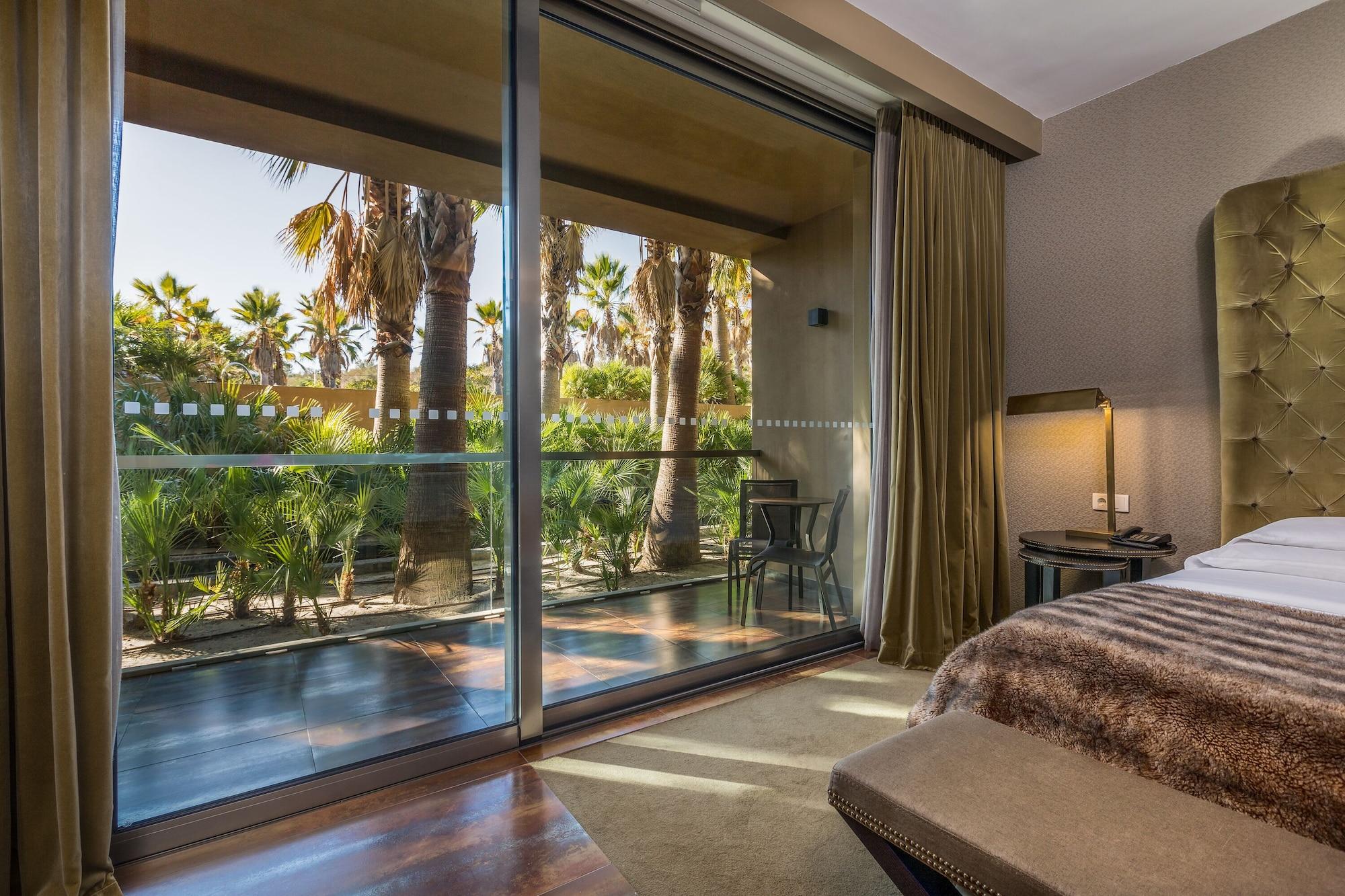Ap Lago Montargil - Conference & Spa Extérieur photo The photo shows a bedroom with large glass doors that lead out to a balcony or terrace. The view outside features palm trees and lush greenery, indicating a tropical or warm climate. Inside the room, there is a bed, a bedside table with a lamp, and a