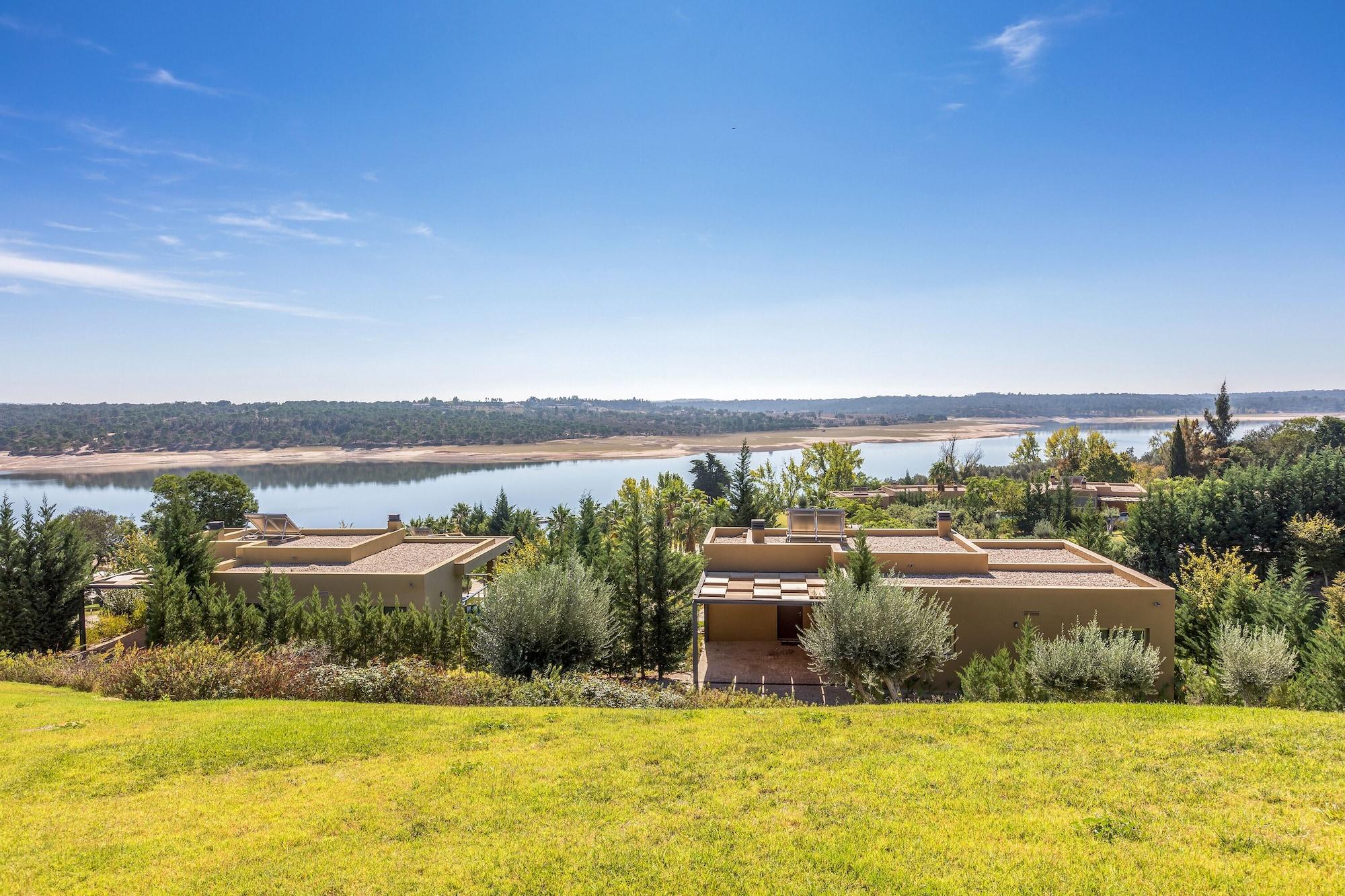 Ap Lago Montargil - Conference & Spa Extérieur photo The photo depicts a scenic landscape featuring a calm body of water, possibly a lake or river, surrounded by lush greenery and trees. In the foreground, there are several modern buildings with flat roofs, set against a vibrant blue sky. The overall s