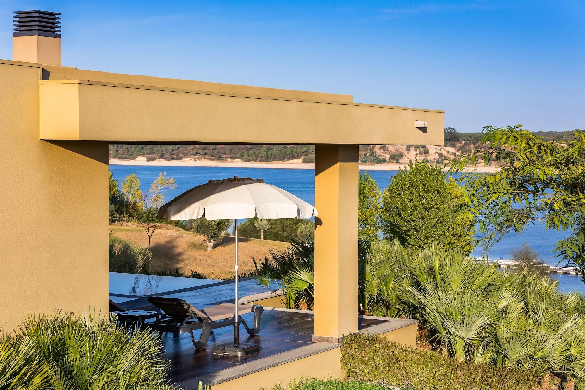 Ap Lago Montargil - Conference & Spa Extérieur photo The photo shows a peaceful outdoor area with a modern architectural structure featuring an overhang. There is a lounge chair positioned beneath a large umbrella, creating a shaded spot to relax. In the background, there is a picturesque view of a bod