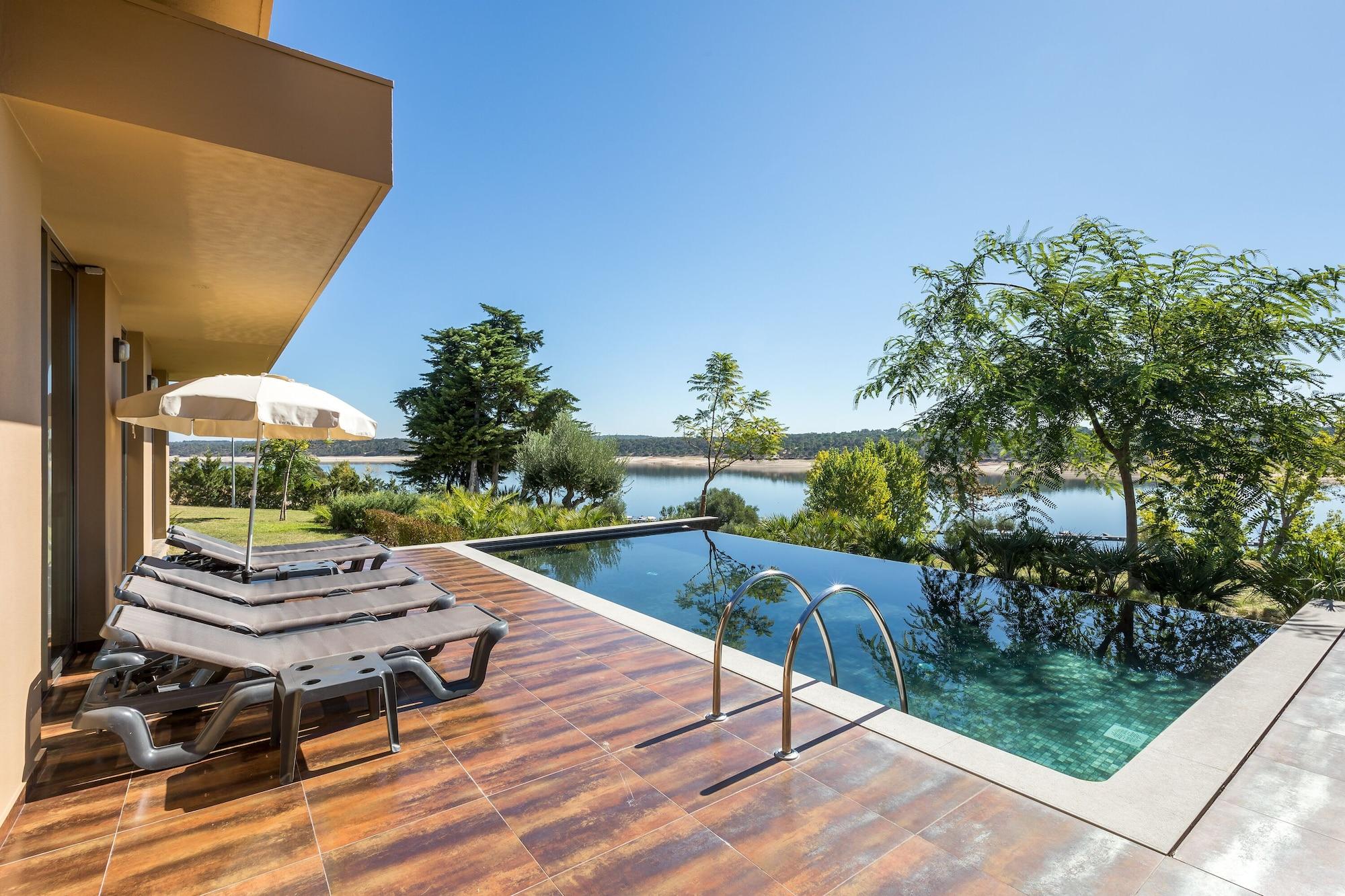 Ap Lago Montargil - Conference & Spa Extérieur photo The photo shows a luxurious outdoor setting by a pool. There are several lounge chairs arranged next to an infinity pool, which overlooks a scenic body of water and lush greenery. A parasol provides shade for relaxation. The sky is clear and blue, in