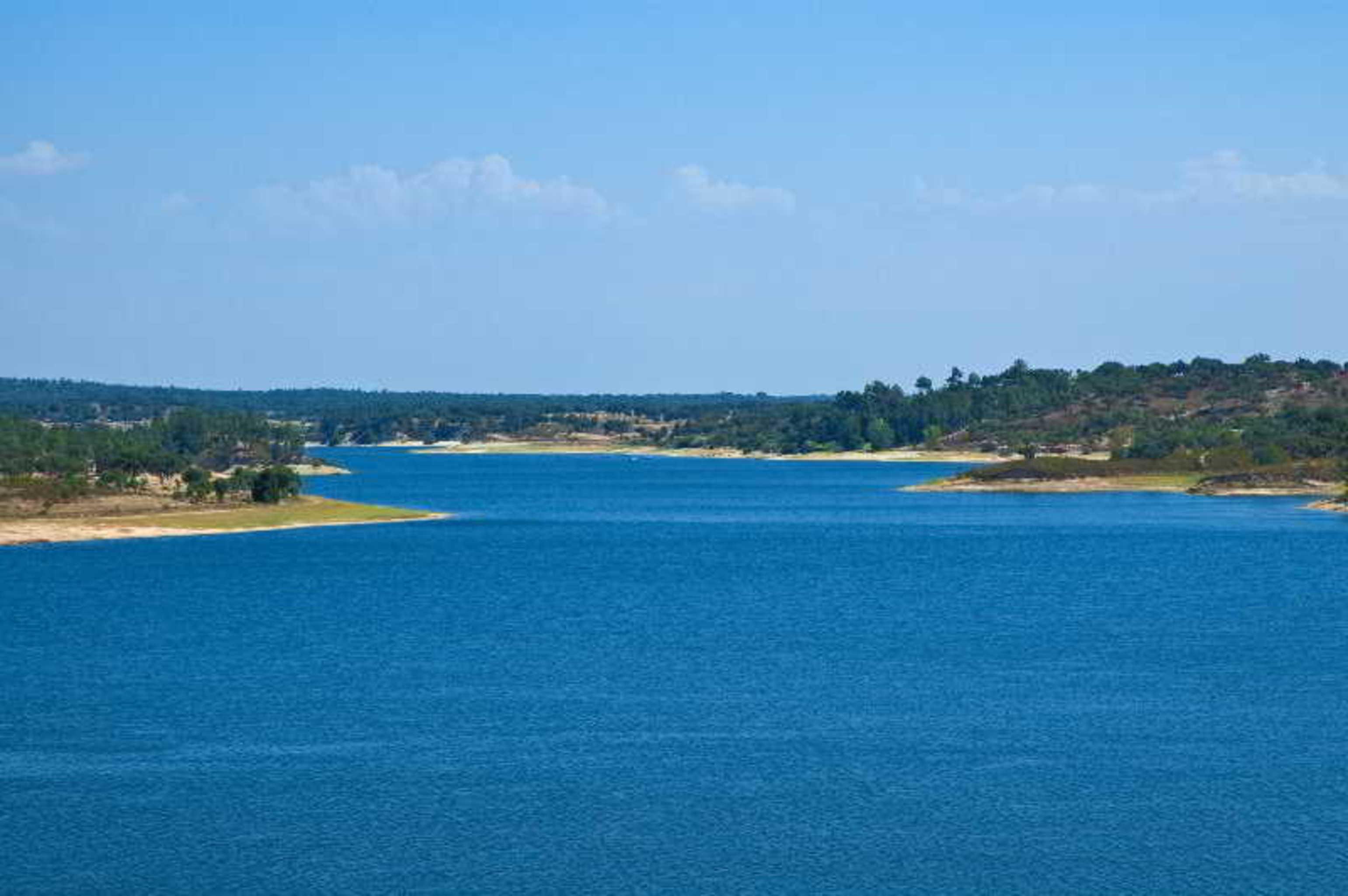 Ap Lago Montargil - Conference & Spa Extérieur photo The photo depicts a serene landscape featuring a large body of water, likely a lake or river. The water appears calm and is reflecting a clear blue sky with some clouds. Surrounding the water, there are patches of green land and trees, indicating a n