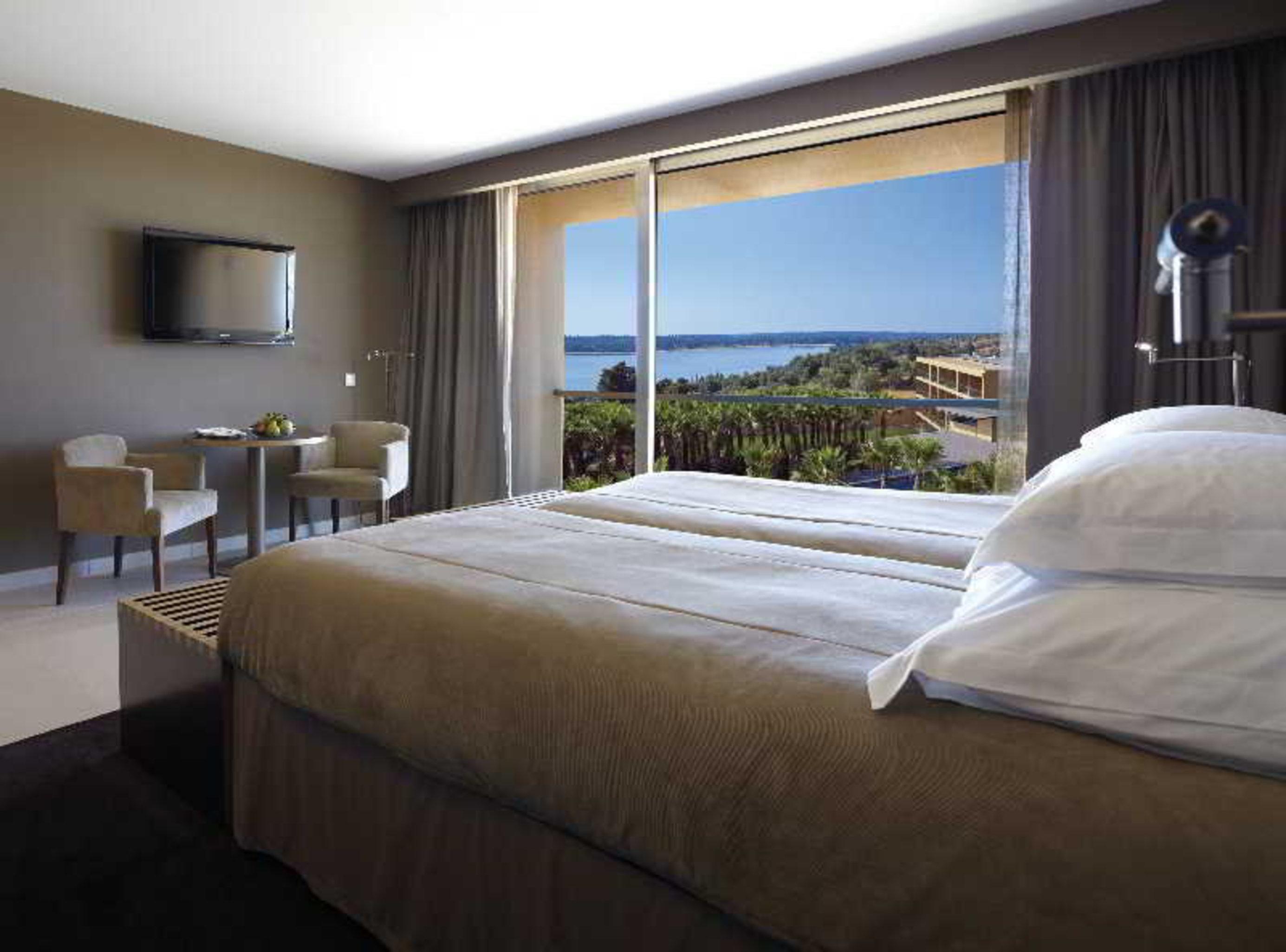 Ap Lago Montargil - Conference & Spa Extérieur photo The photo shows a modern hotel room with a large window offering a view of the sea and mountains. The room features a comfortable bed with white pillows and brown bedding. There is a small sitting area with two chairs and a table, as well as a wall-m