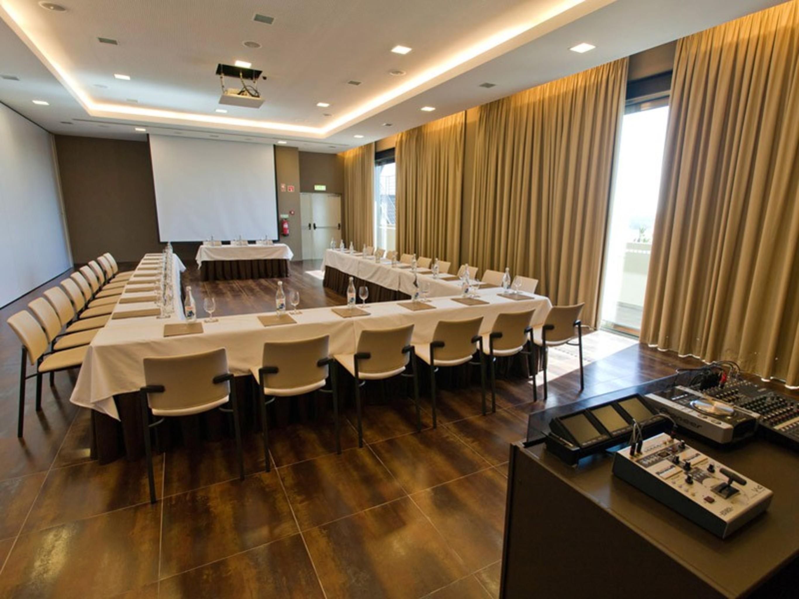 Ap Lago Montargil - Conference & Spa Extérieur photo The photo shows a modern conference room set up for a meeting. There are several tables arranged in a U-shape, surrounded by chairs. A large screen and projector are positioned at the front, with a table for speakers or presenters. The room has large