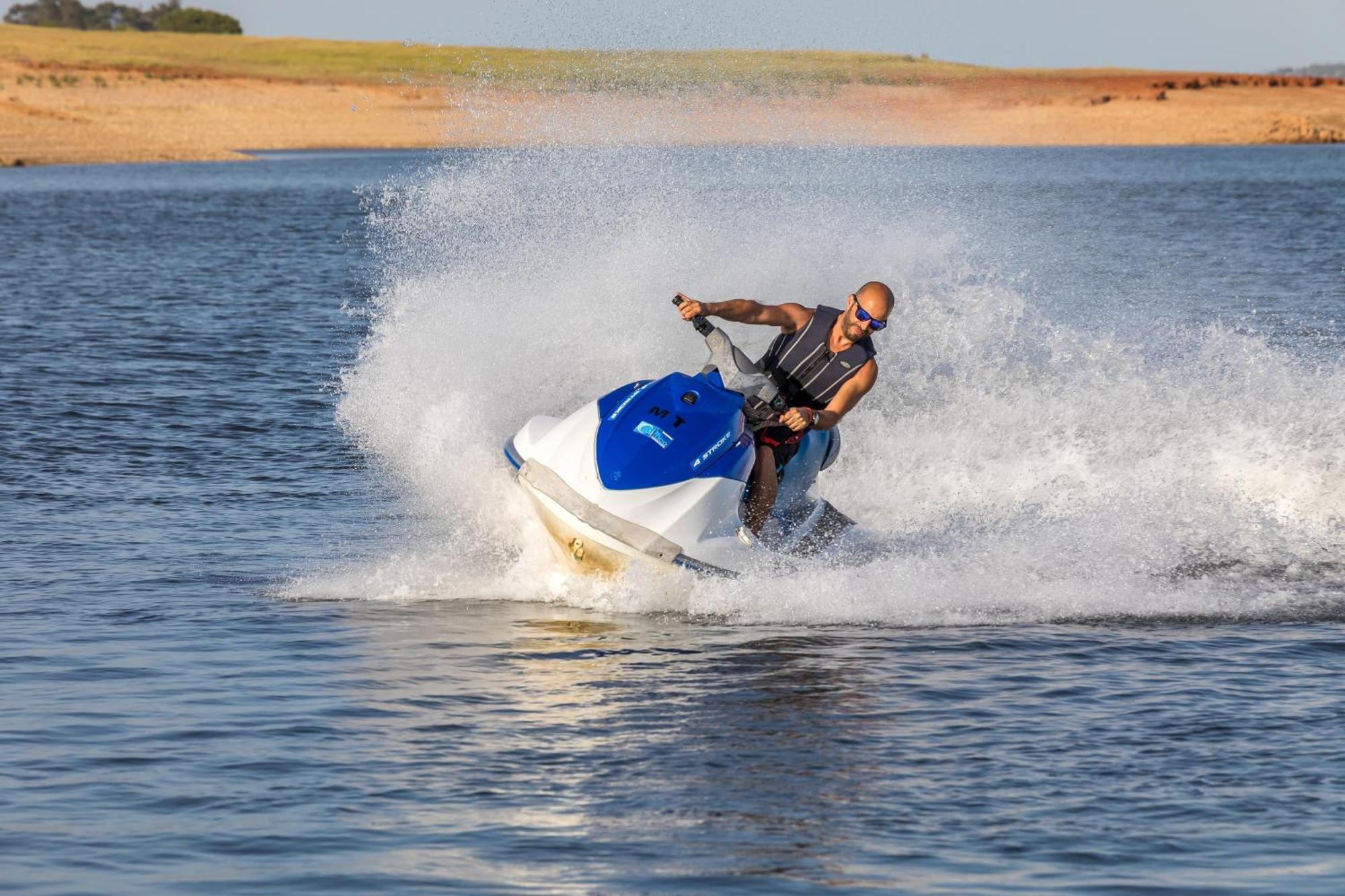 Ap Lago Montargil - Conference & Spa Extérieur photo The photo shows a person riding a jet ski over a body of water. The rider is wearing sunglasses and a life vest, leaning forward as the jet ski accelerates, creating a spray of water around them. The background features a natural landscape, likely a 