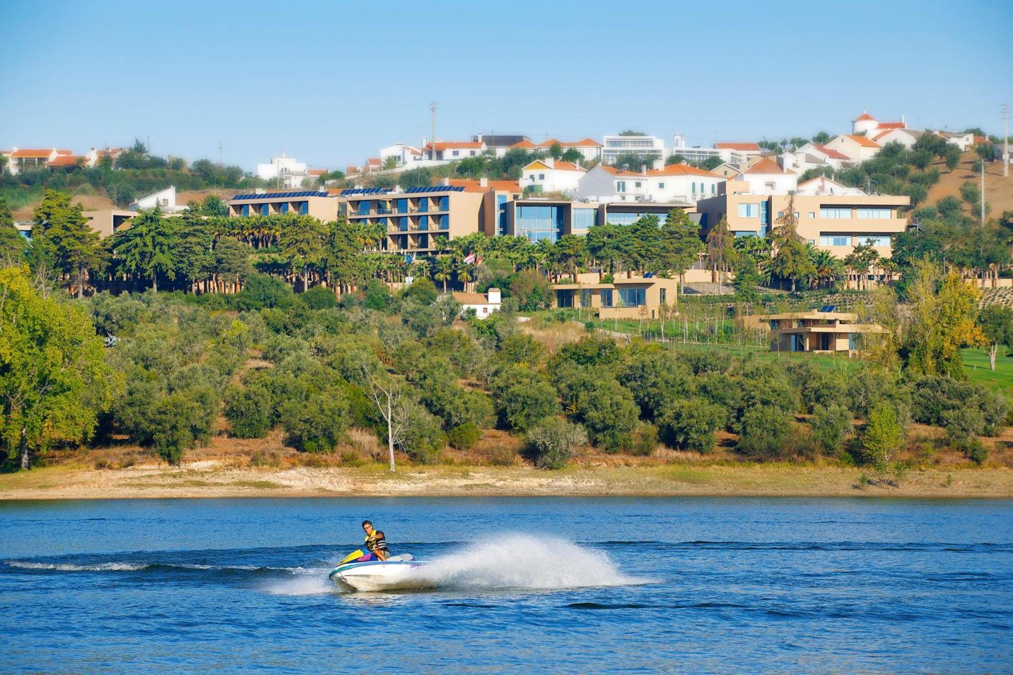 Ap Lago Montargil - Conference & Spa Extérieur photo The photo shows a person riding a jet ski on a body of water, creating a spray as they speed across the surface. In the background, there are modern buildings and a hillside with trees and greenery. The sky is clear and blue, suggesting a bright and 