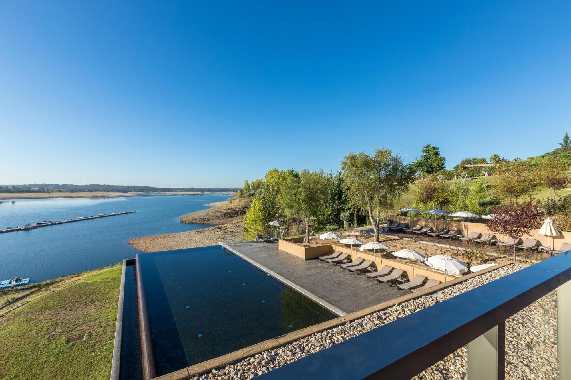 Ap Lago Montargil - Conference & Spa Extérieur photo The photograph depicts a scenic view of a tranquil water body, likely a lake or river, bordered by lush greenery. In the foreground, there is an infinity pool that blends seamlessly with the water beyond. Comfortable lounge chairs are arranged on a w