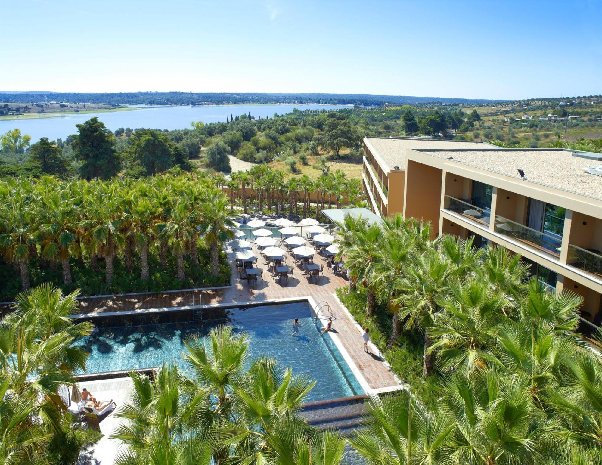 Ap Lago Montargil - Conference & Spa Extérieur photo The photo shows a scenic view of a modern hotel or resort. In the foreground, there is a swimming pool surrounded by palm trees, with lounge chairs positioned beside it. Sun umbrellas provide shade over the chairs. The background features a lush land