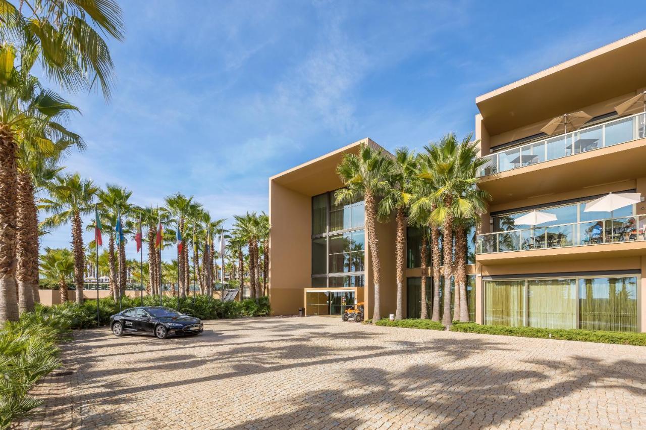 Ap Lago Montargil - Conference & Spa Extérieur photo The photo shows a modern building with a sleek design, featuring large glass windows and balconies. The structure is surrounded by a well-maintained outdoor area with palm trees, adding a tropical feel. In front of the building, there is a paved park