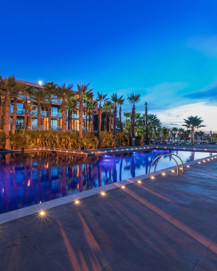 Ap Lago Montargil - Conference & Spa Extérieur photo The image shows a serene and inviting outdoor area with a large swimming pool that features a beautiful blue hue, likely illuminated by lights. Surrounding the pool are well-maintained palm trees and lush greenery, creating a tropical atmosphere. In 